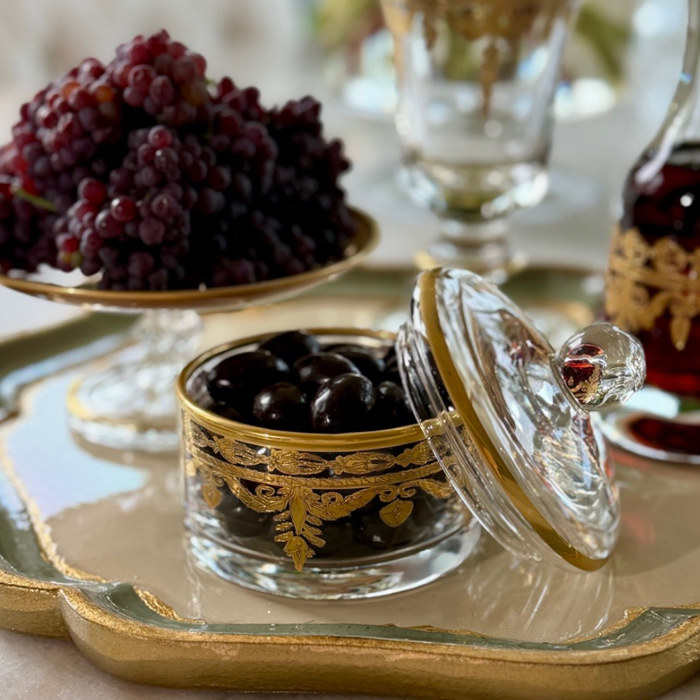 A Vetro gold small covered dish filled with chocolate almonds on a Florentino tray with grapes and wine in background