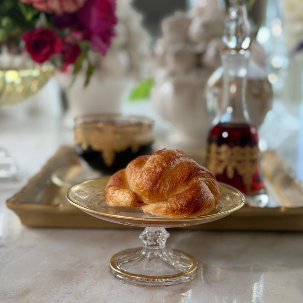 A croissant sitting on a Vetro gold small patisserie dish with pedestal on a table with coffee and other vetro gold items