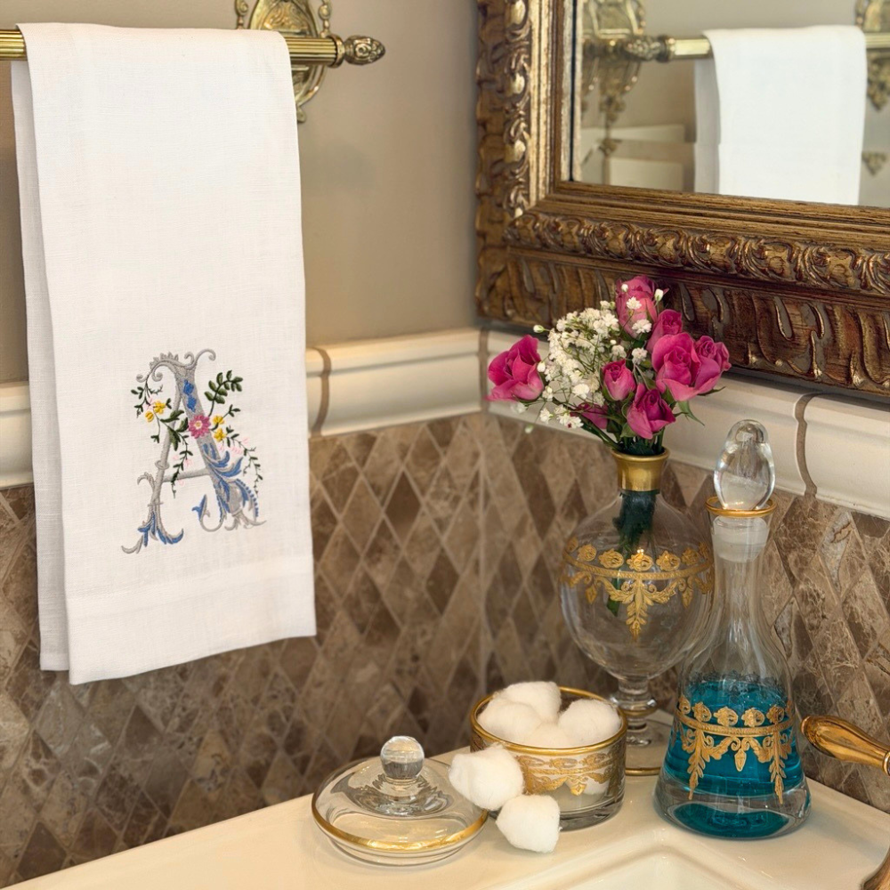 A bathroom scene with a Heritage floral Monogram Linen towel, vetro gold covered dish with cotton balls and Small Decanter with mouthwash next to a sink