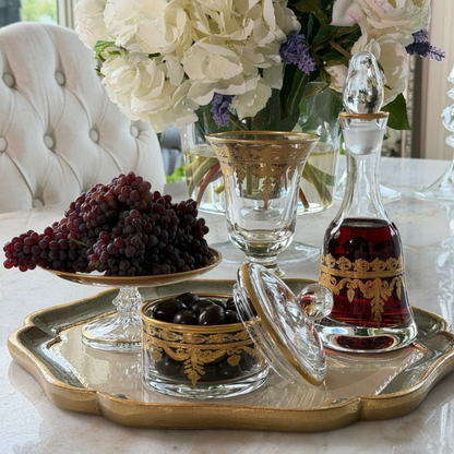 A florentino wooden tray holding Vetro gold small decanter, patisserie dish, small covered dish and a wine glass on a table with flowers