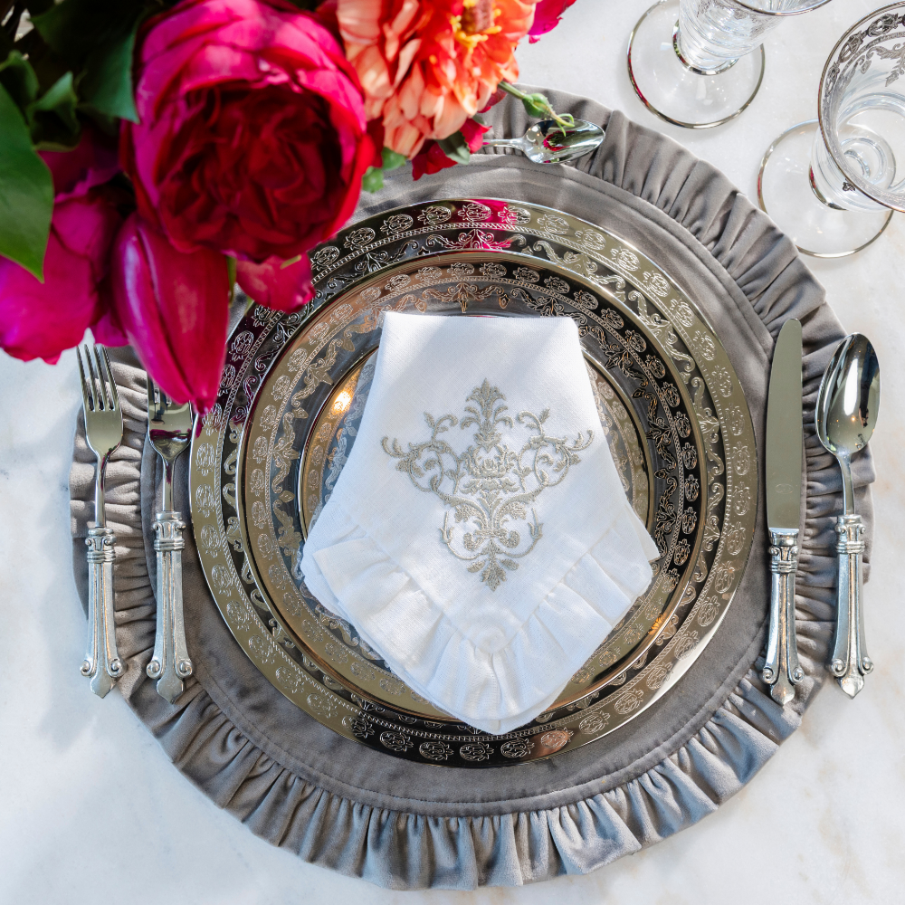 Elegant place setting with Vetro Platinum plates on a grey velvet ruffle placemat with a white linen victorian linen napkin with ruffle folded on it