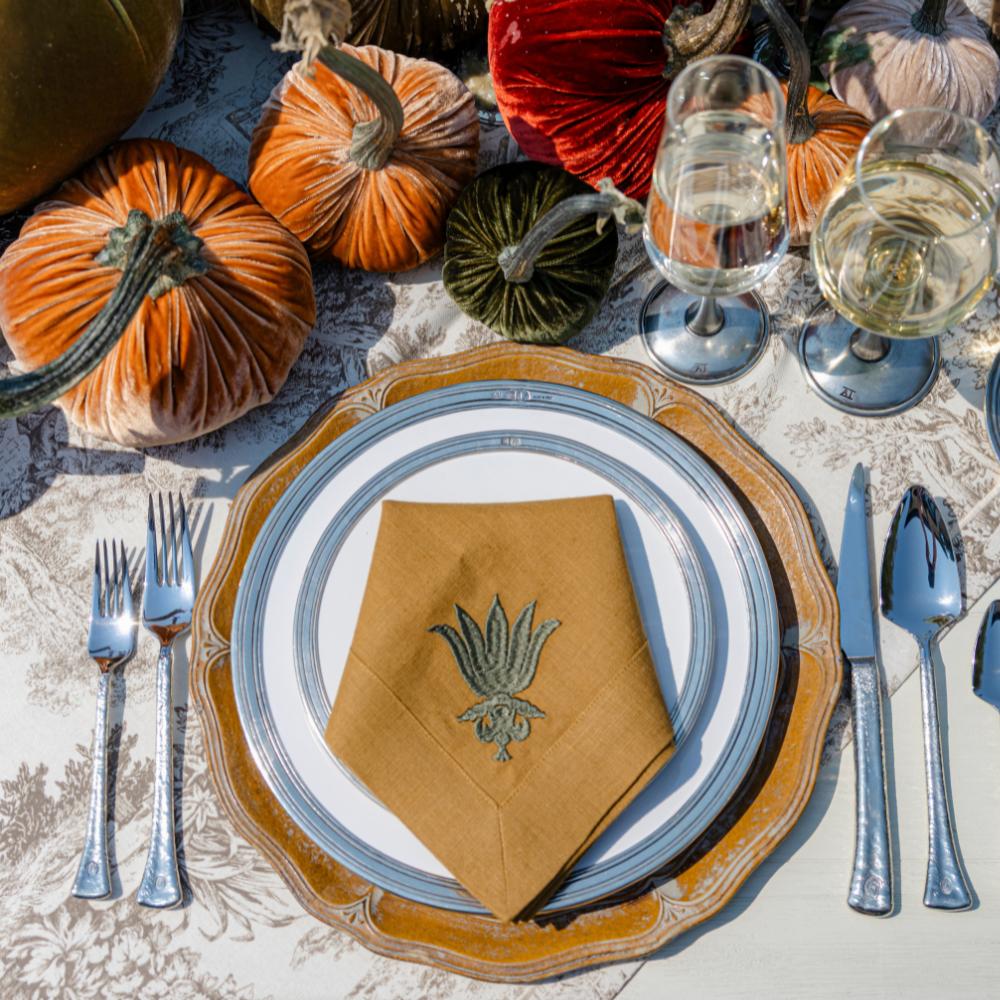 A Tuscan place setting on a fall table with velvet pumpkins and Siena wine glasses with a villa floret embroidered large camel linen napkin folded and placed on plate