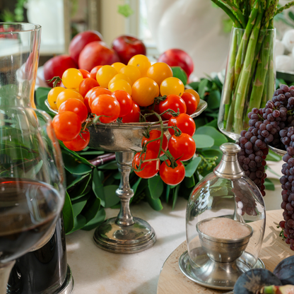 A vintage round petite pewter pedestal bowl filled will small tomatoes that is part of a charcuterie spread on a table with asparagus, grapes and wine