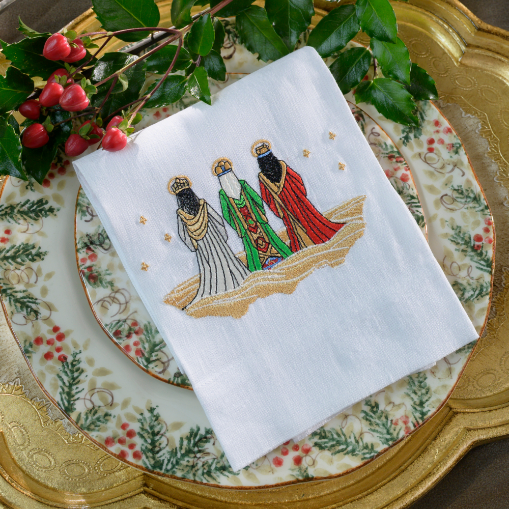 White linen tea towel embroidered with a three wisemen design folded on a natale plate with greenery and gold tray background