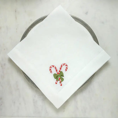White linen Candy Cane Lane napkin with red and green embroidered candy cane motif, laid flat on a marble surface