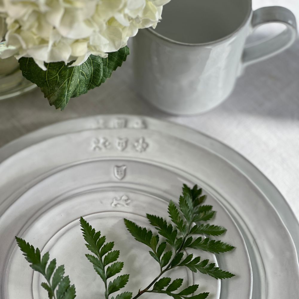 White Firenze Stoneware plate with embossed stamp design, green leaves, and a mug on a light surface.