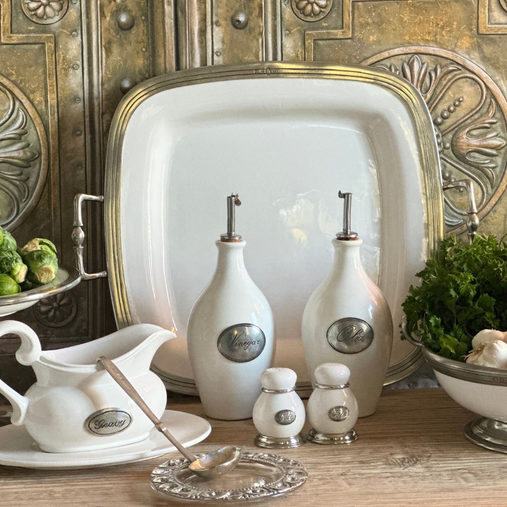 Tuscan white ceramic and pewter serving pieces including oil and vinegar bottles, salt and pepper shakers, and a platter against a metallic wall.
