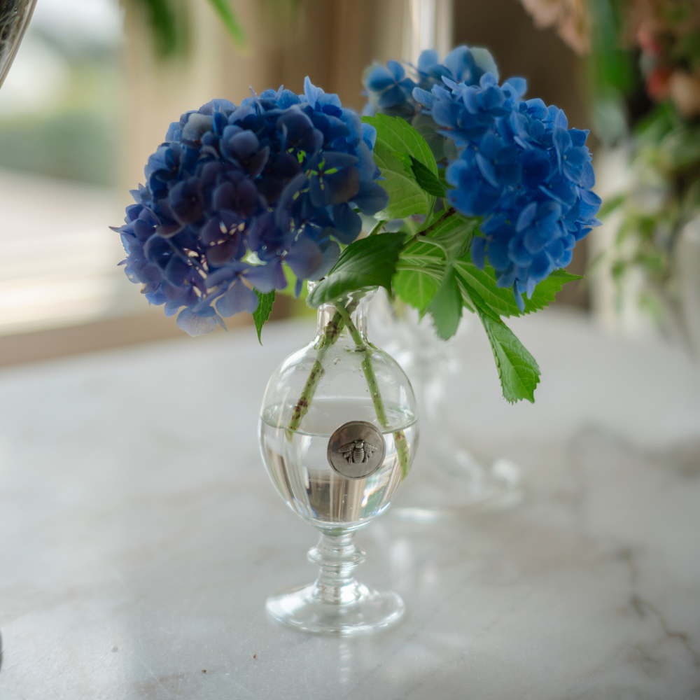 Glass bud vase with pewter tab with engraved bee on a white table holding blue hydrangeas