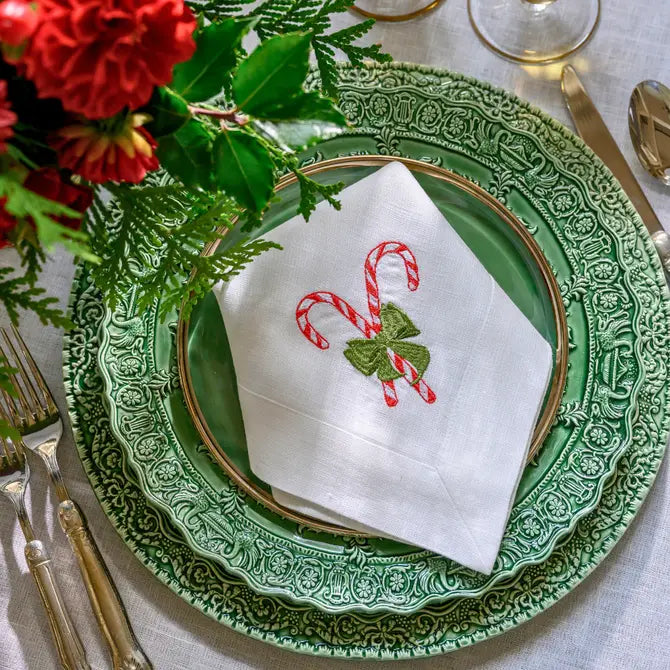 White linen napkin with embroidered candy cane and green bow motif, folded on a green renaissance ceramic charger with hotel collection flatware and red floral centerpiece