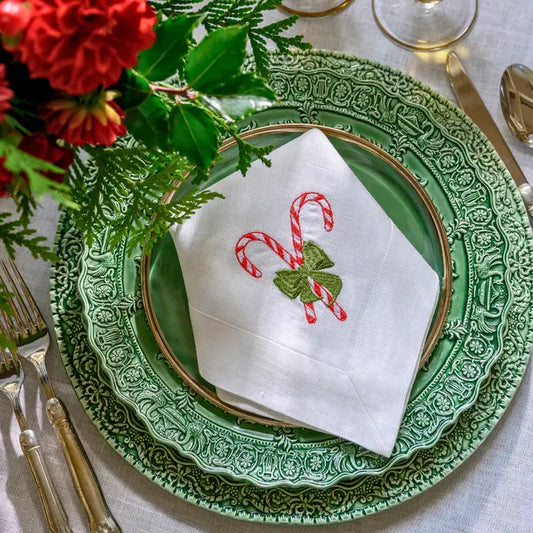 White linen napkin with embroidered candy cane and green bow motif, folded on a green renaissance ceramic charger with hotel collection flatware and red floral centerpiece