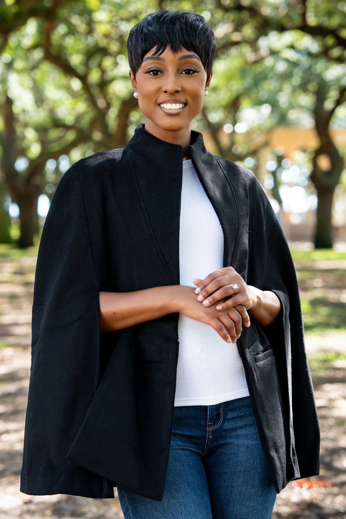 Woman wearing a black cape over a white shirt and blue jeans, standing outdoors with trees in the background.
