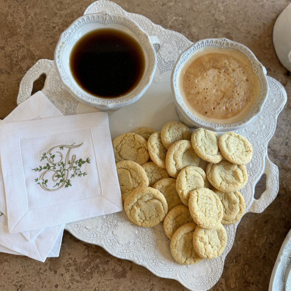A Merletto antique white square platter with handles on a table with 2 mugs with coffee and sugar cookies on it with monogram linen napkins.