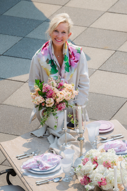 Woman wearing the Portofino Summer Garden Silk Scarf placing pink flowers on a table on a patio
