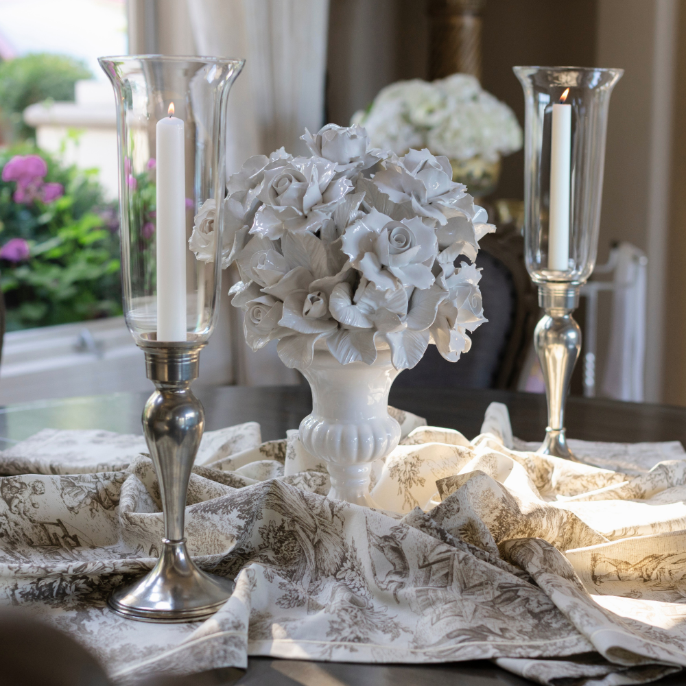 A decorative tabletop arrangement featuring a white floral ceramic centerpiece in a white urn, flanked by two tall giovanna clear glass tapered hurricane candle holders with white candles, set on draped Montefalco Latte Toile patterned fabric.