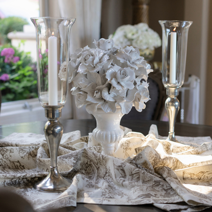 A decorative tabletop arrangement featuring a white floral ceramic centerpiece in a white urn, flanked by two tall giovanna clear glass tapered hurricane candle holders with white candles, set on draped Montefalco Latte Toile patterned fabric.