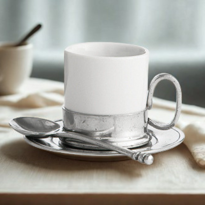 An ceramic Tuscan espresso cup with pewter bottom and handle, sitting on a pewter saucer with a spoon on a table