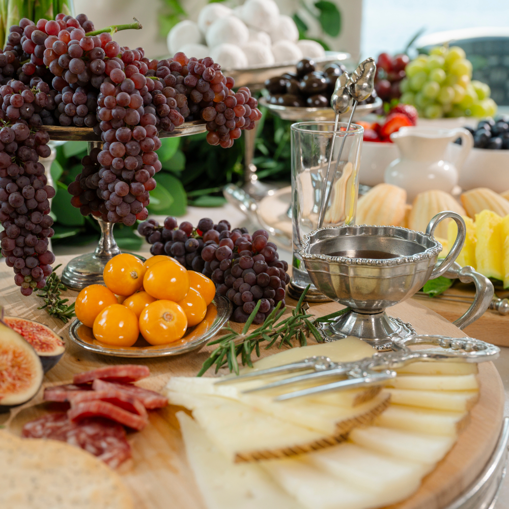 A vintage beaded pewter piccolo creamer filled with honey on a charcuterie board with cheese and grapes and a bread fork